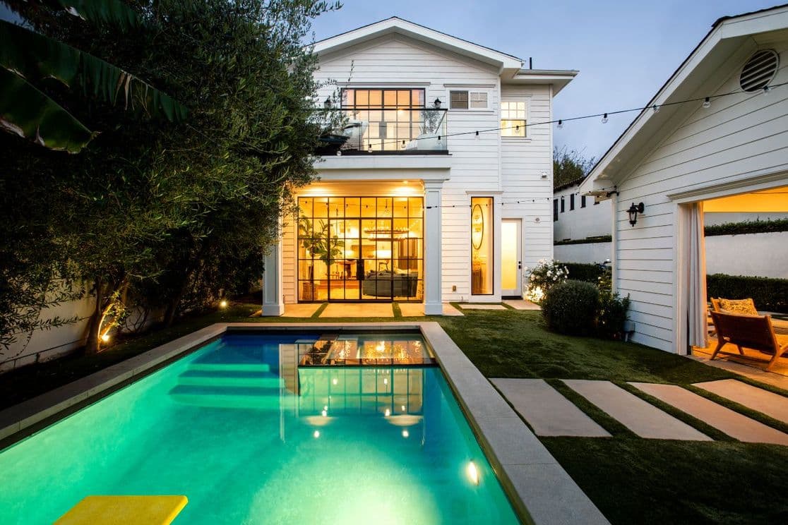 Modern white house with large windows, patio lights, and a lit swimming pool in the backyard. Adjacent garage and lush greenery. Evening setting.