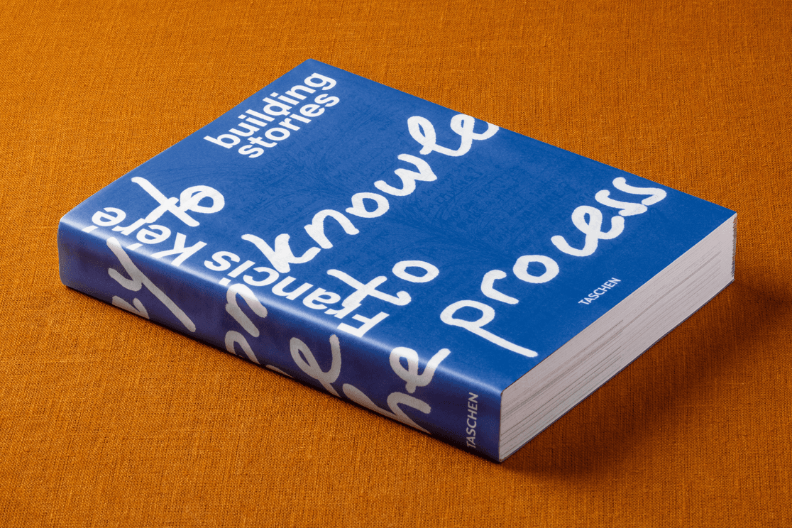 Ein dickes blaues Buch mit weißem, handschriftlich anmutendem Text auf dem Einband, das auf einer orangefarbenen, strukturierten Stoffoberfläche liegt.