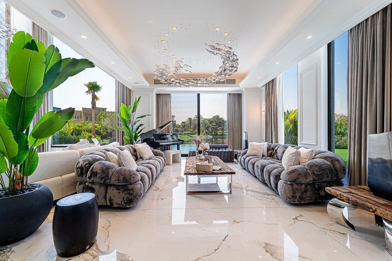 Luxurious living room with plush sofas, a grand piano, large windows, marble floor, and a unique chandelier, overlooking a garden view.