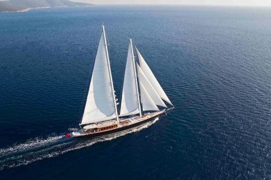 Aerial view of a sailing yacht with white sails cutting through calm blue ocean water under a clear sky. Land is visible in the distant background.