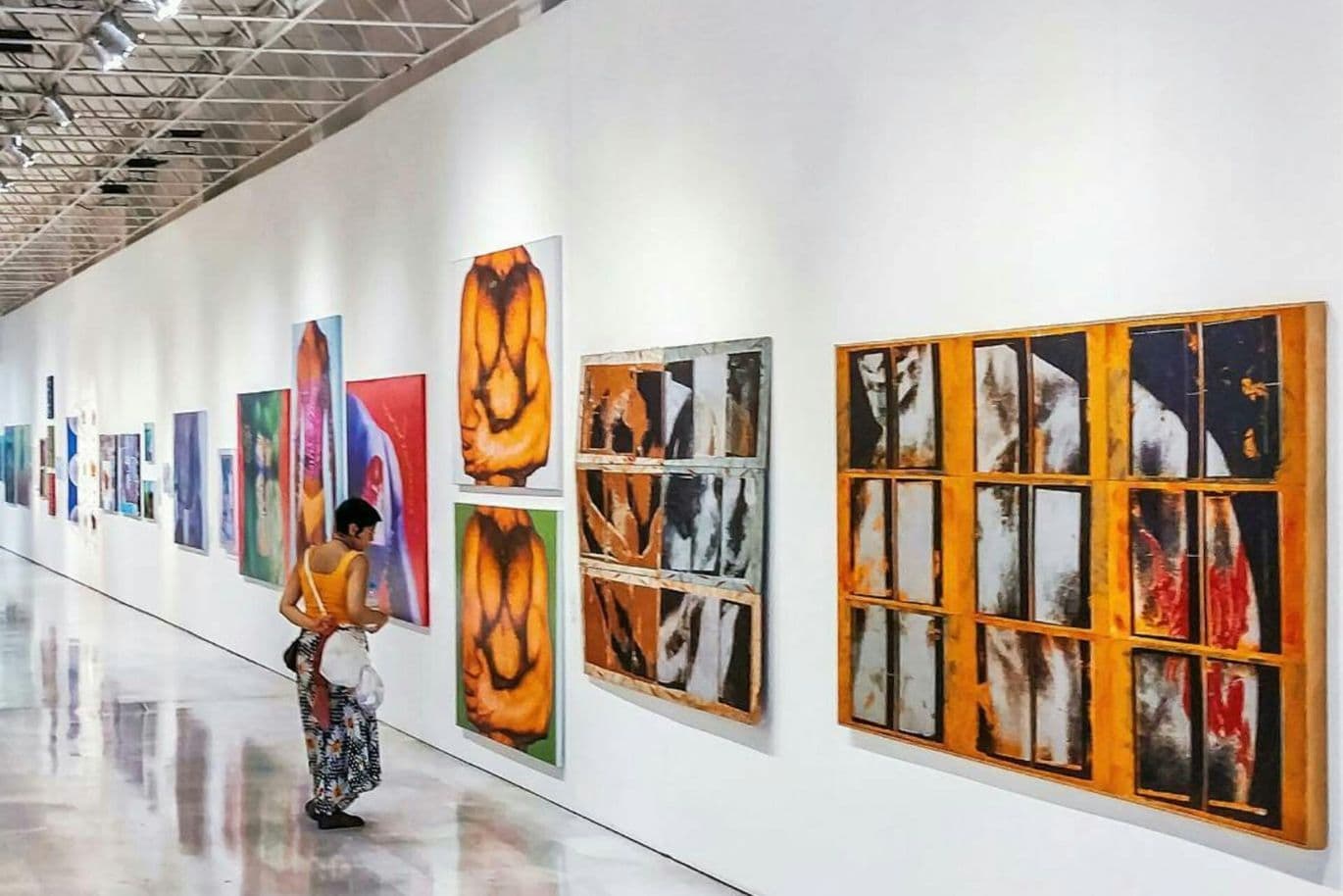 A person in a yellow top and patterned skirt observes colorful abstract paintings on a long white gallery wall.