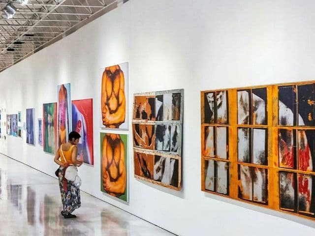 A person in a yellow top and patterned skirt observes colorful abstract paintings on a long white gallery wall.
