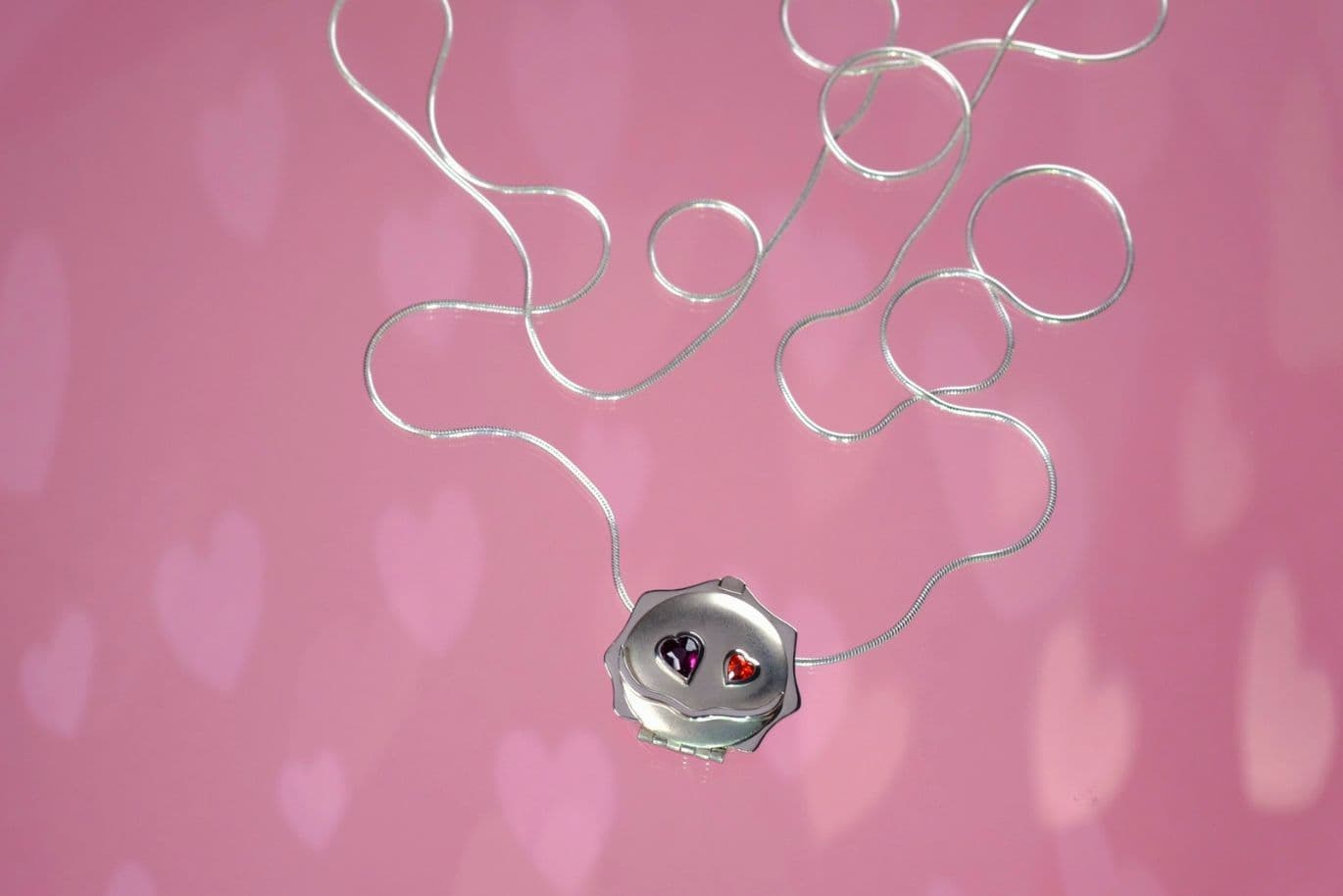 Eine silberne Halskette mit einem Medaillon, der mit zwei Edelsteinen besetzt ist, vor einem rosa Hintergrund mit Herzmuster.