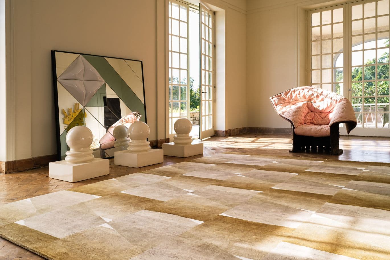 Sunlit room with geometric mirrors, abstract sculptures, and a unique pink chair. Large windows reveal a lush garden view.