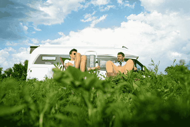 Two people relax in front of a white camper van on a grassy field under a partly cloudy sky.