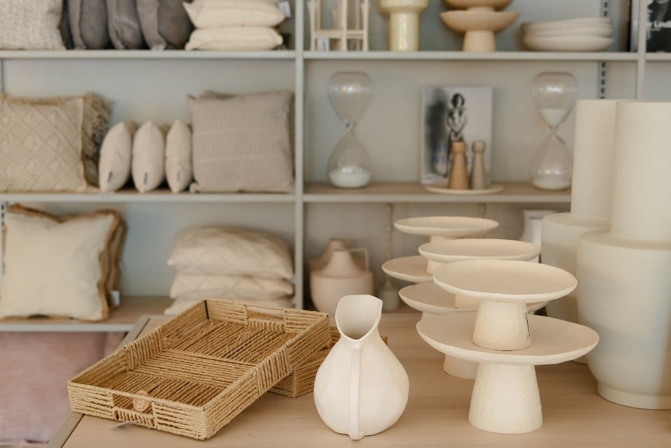An exhibition of interior design items in neutral colors, including cushions, woven trays, vases, and hourglasses, arranged on shelves and a table.
