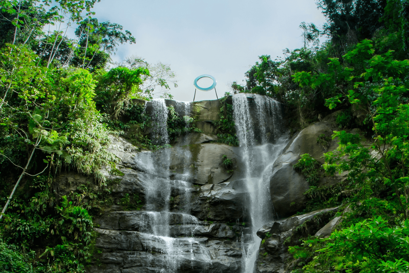Ein Wasserfall, der über eine felsige Klippe hinabstürzt, umgeben von üppigem Grün, wobei oben ein großes rundes Bauwerk zu sehen ist.
