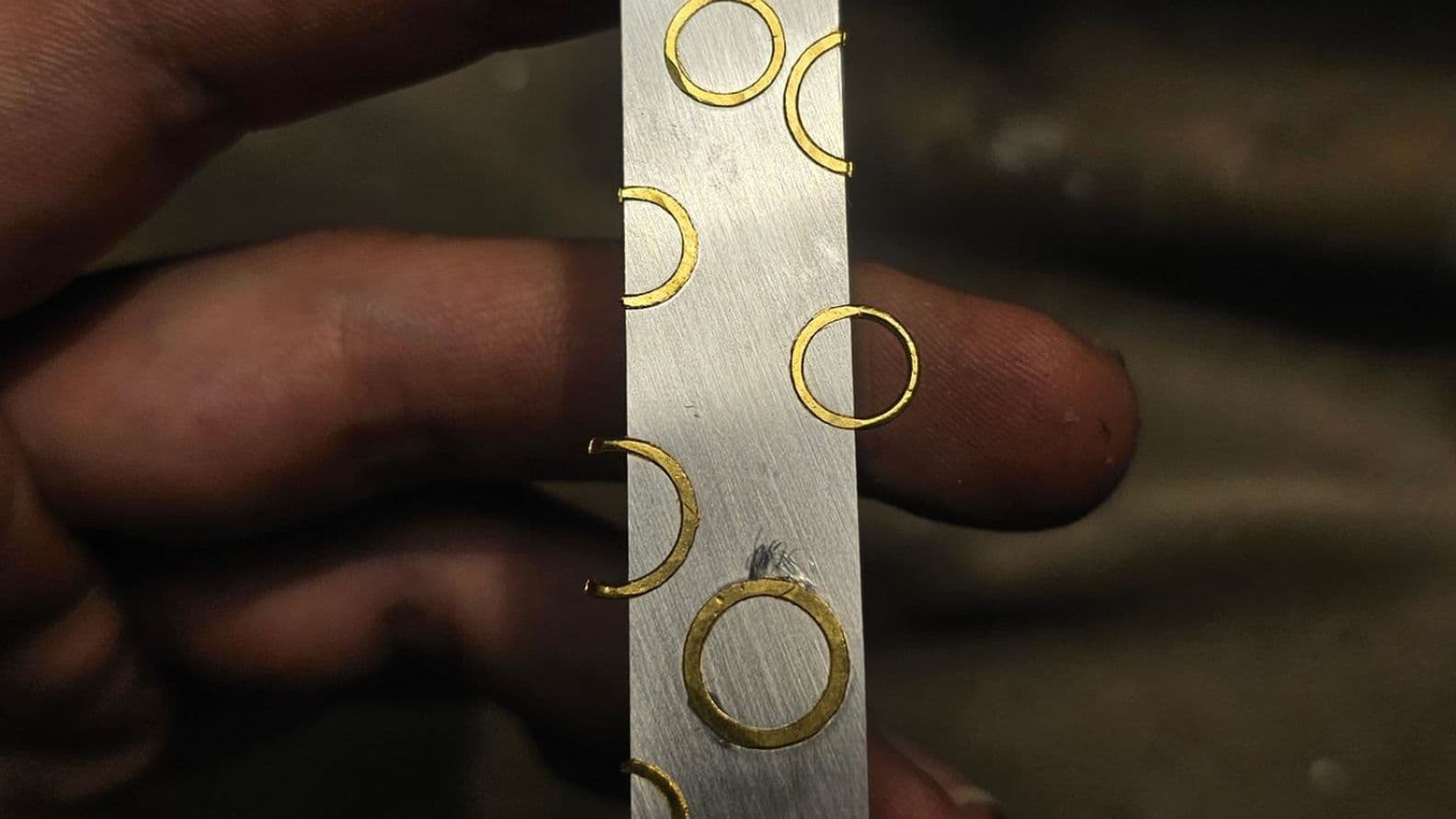 Finger, die eine Lippenstifthülle aus Metall mit mehreren kleinen Goldringen halten, vor einem unscharfen Hintergrund.