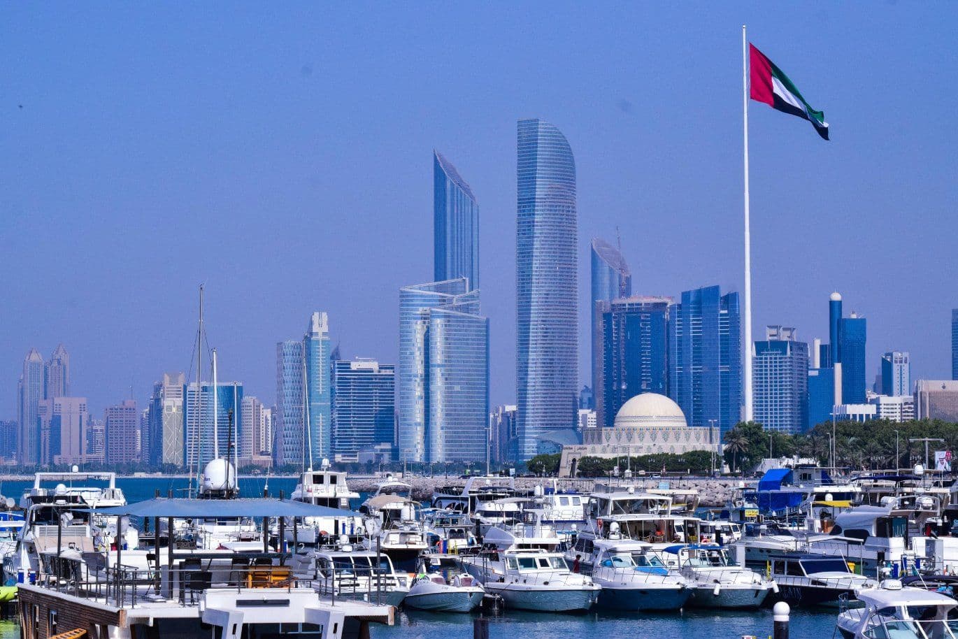 Skyline of Abu Dhabi with modern skyscrapers, a large UAE flag, and numerous boats docked in a marina under a clear blue sky.