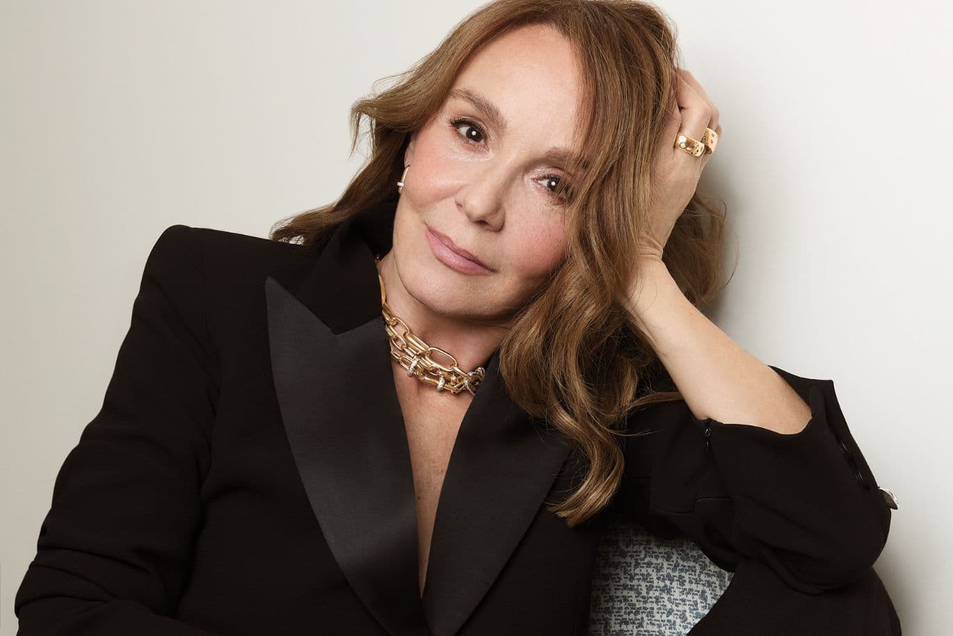 Portrait of a woman with light brown hair in a black blazer, leaning against a wall with one hand in her hair, wearing gold jewelry.