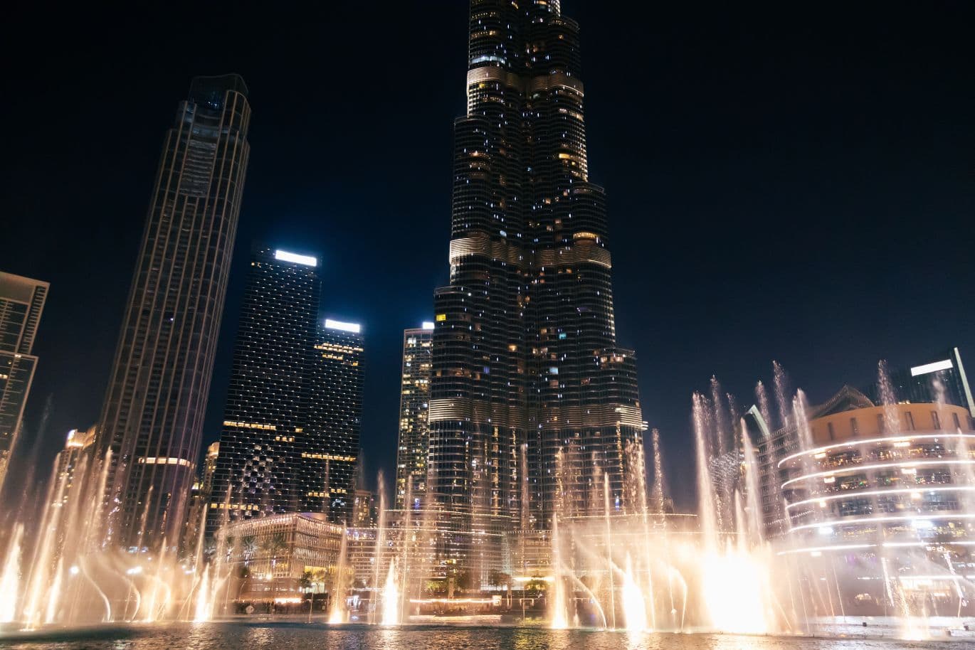 Night view of a cityscape with illuminated skyscrapers and a fountain display in the foreground, set against a dark sky.