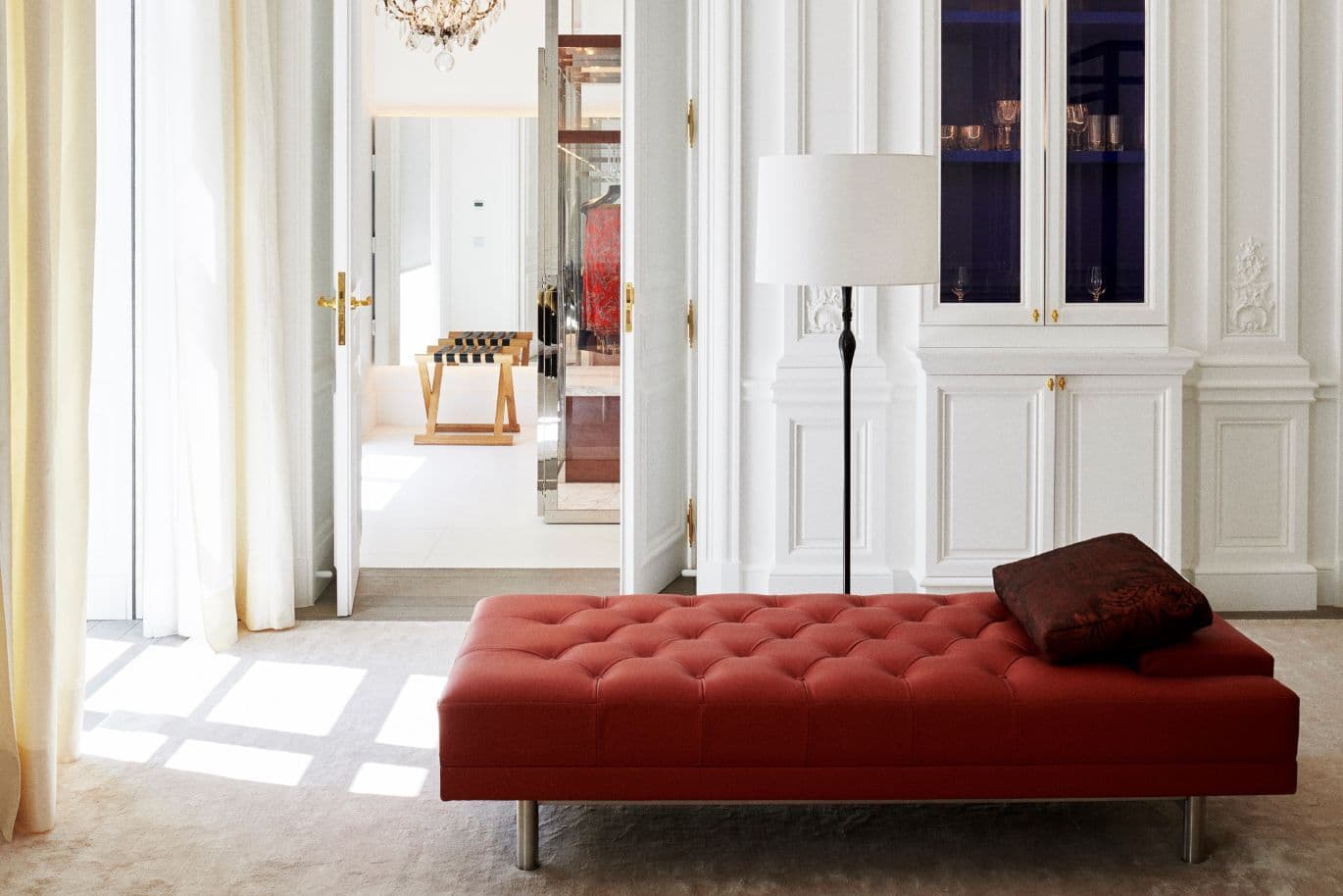 Elegant room with a red chaise longue with button detailing, floor lamp and sunlight filtering through curtains. Open doors lead to a bright adjoining room.