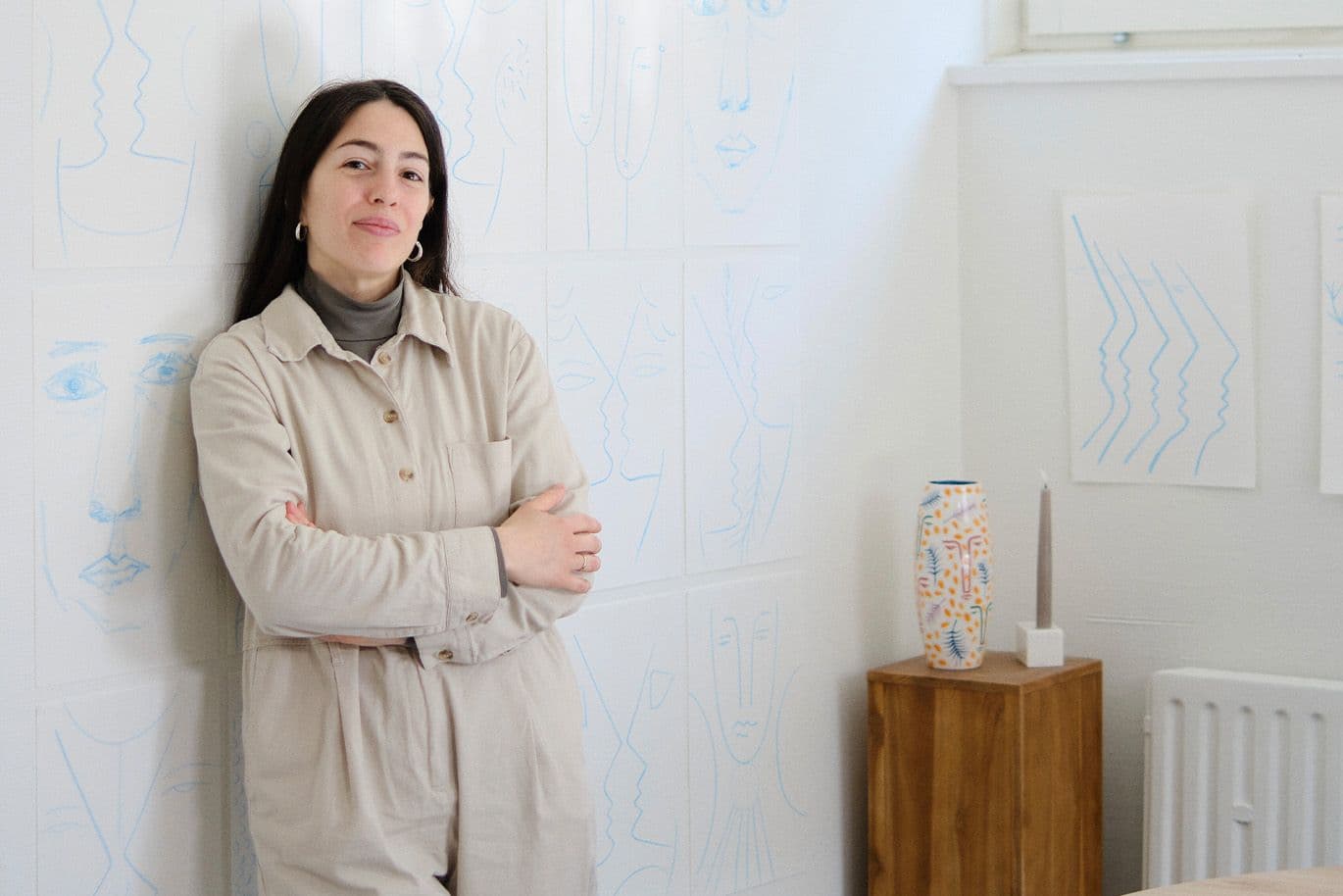 Γυναίκα με μπεζ φόρμα στηρίζεται σε τοίχο με καλλιτεχνικά σκίτσα σε μπλε γραμμές.