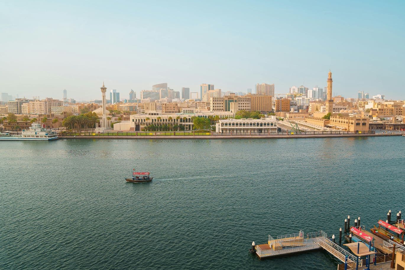 A boat on a wide river with Dubai in the background, featuring modern and traditional buildings under a clear blue sky.