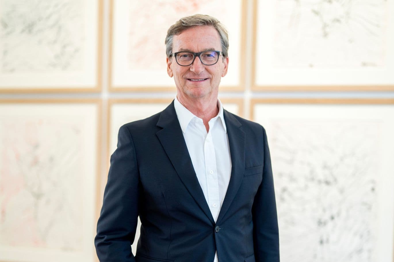 Porträt eines Manns mit Brille im dunklen Anzug, lächelnd vor Bildern in heller schlichter Galerie.