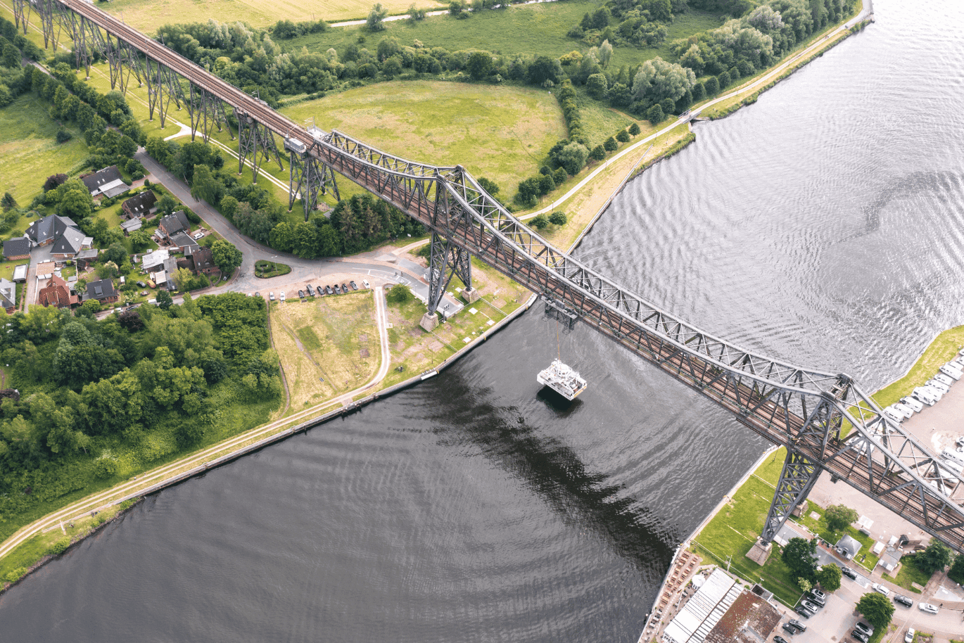Aerial view of a large steel bridge over a river, with a ferry passing underneath, surrounded by green fields and residential areas.