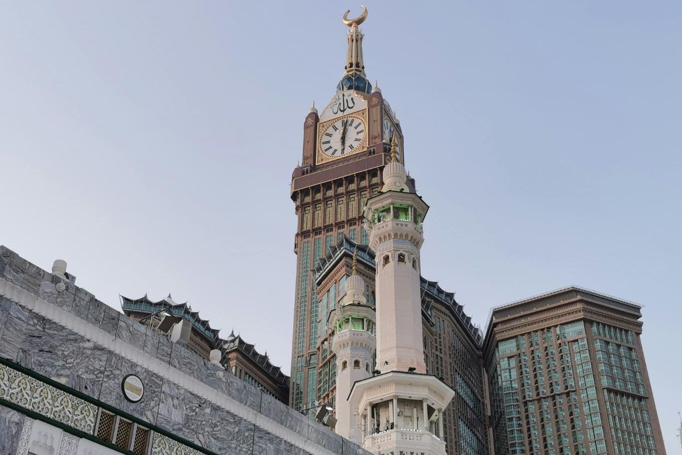 Majestic tower with a large clock face and crescent at the top, surrounded by intricate buildings, against a clear blue sky.