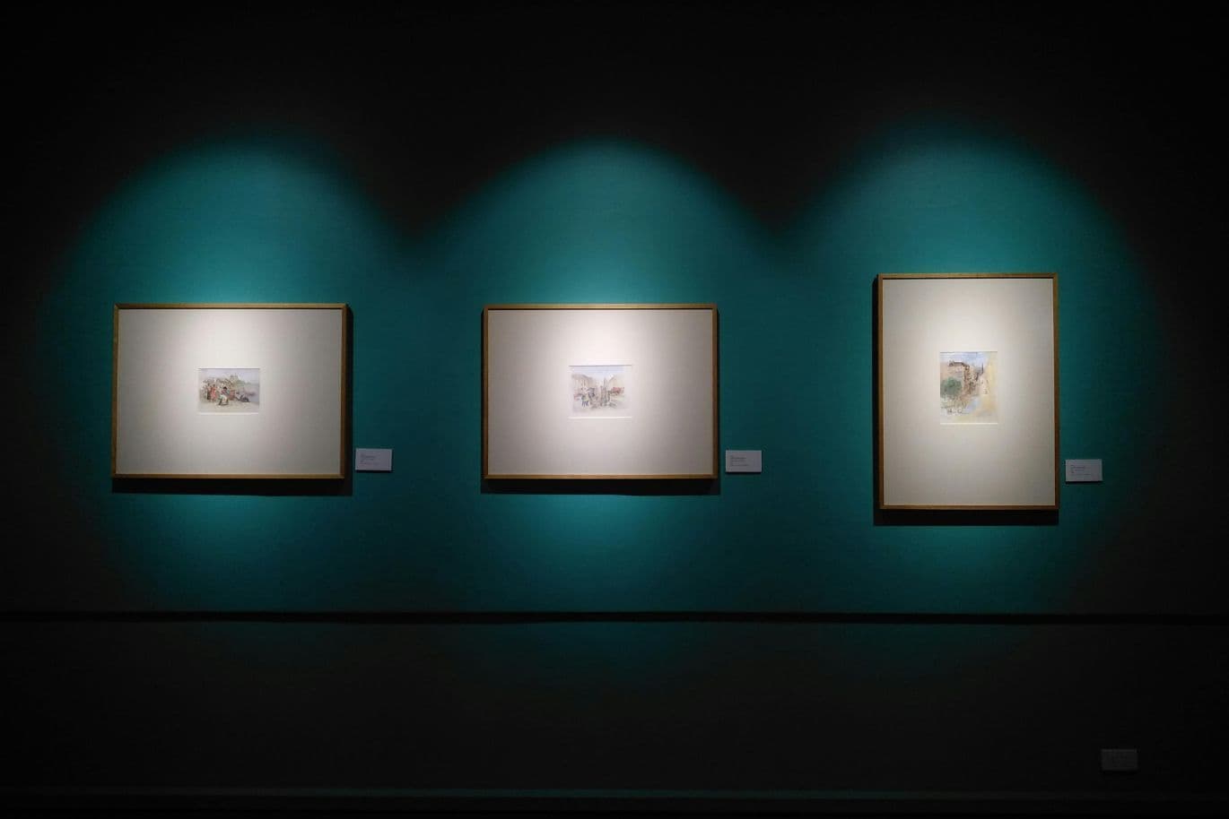 Three framed artworks illuminated on a teal wall in a dimly lit gallery, each with a small label beside it.