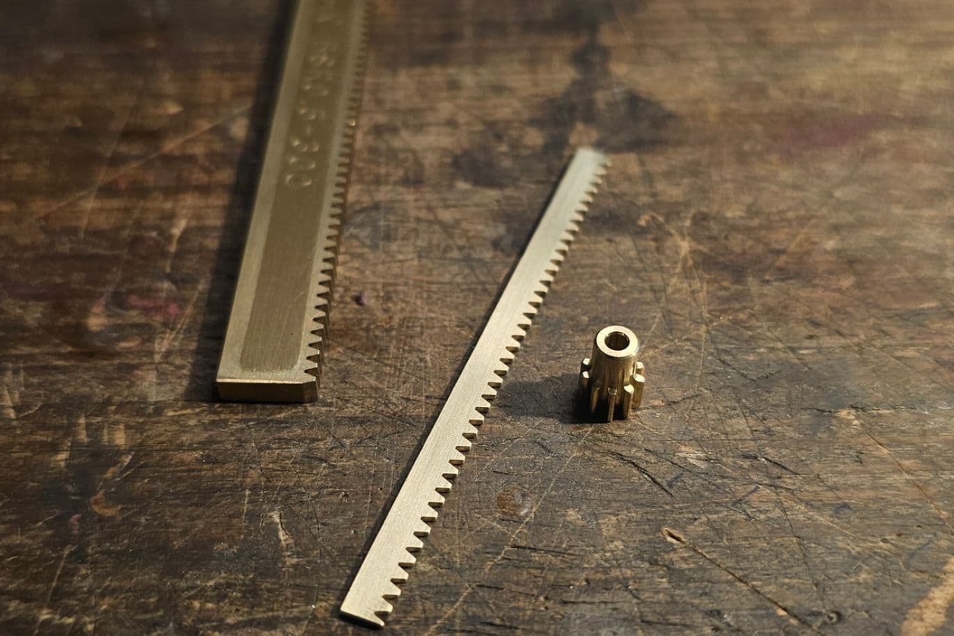 Nahaufnahme von Goldschmiede-Werkzeugen, darunter eine Zahnstange, eine kleinere Stange mit Zähnen und ein zylindrisches Zahnrad, auf einer Holzoberfläche.