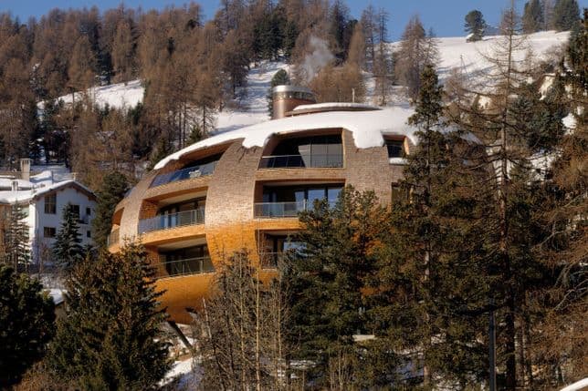 Unique, spherical wooden house with large windows, nestled in a snowy, forested hillside, surrounded by bare winter trees.