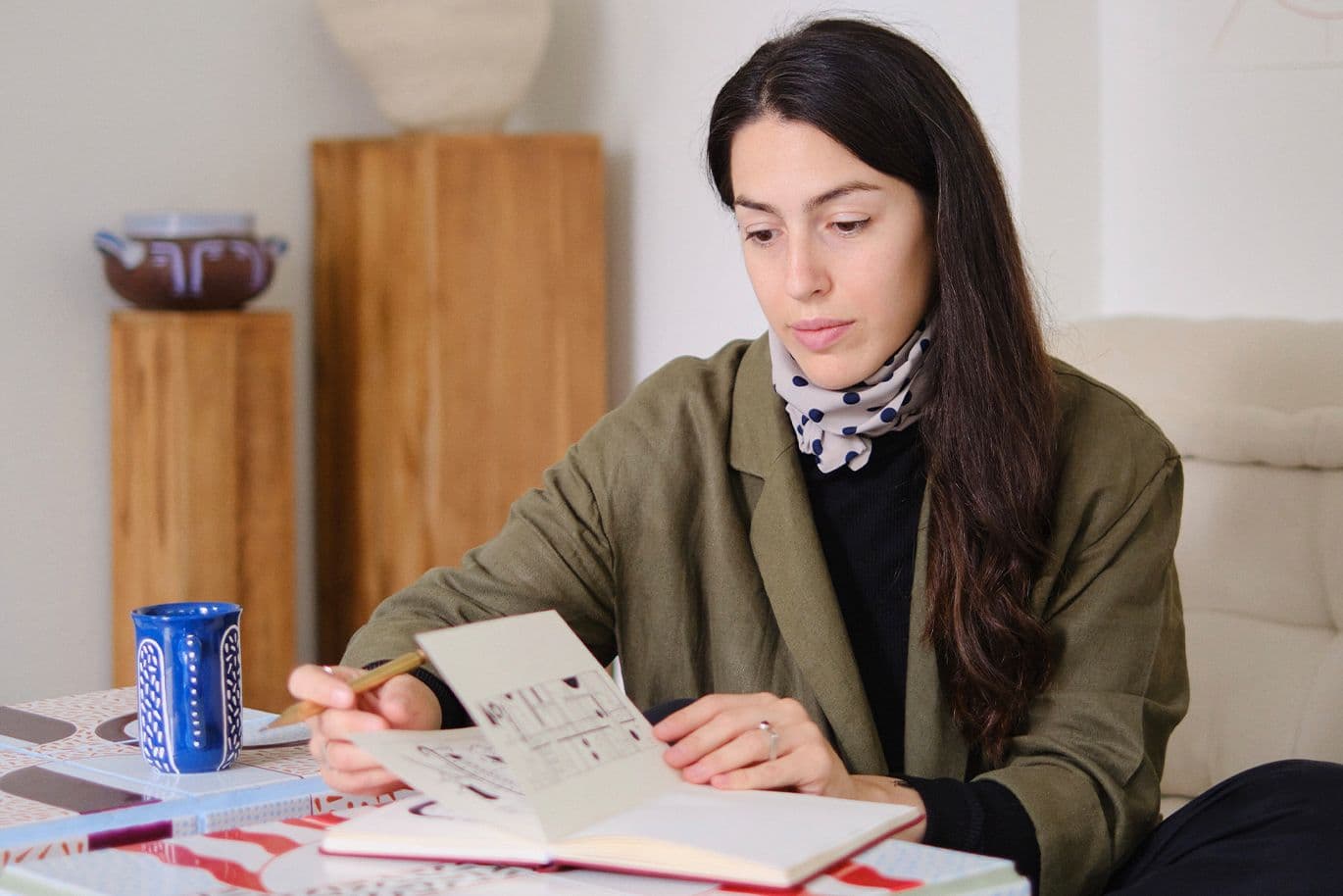 Γυναίκα κάθεται σε τραπέζι, κρατά σημειωματάριο και μολύβι, δίπλα σε μπλε κούπα.