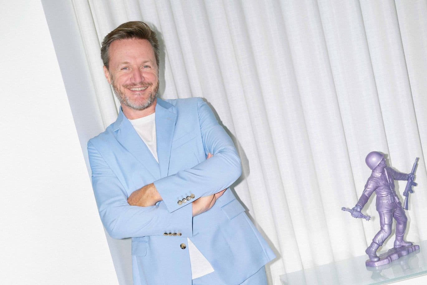 Person in a light blue suit, smiling with arms crossed, stands beside a small purple astronaut sculpture. White curtains in the background.