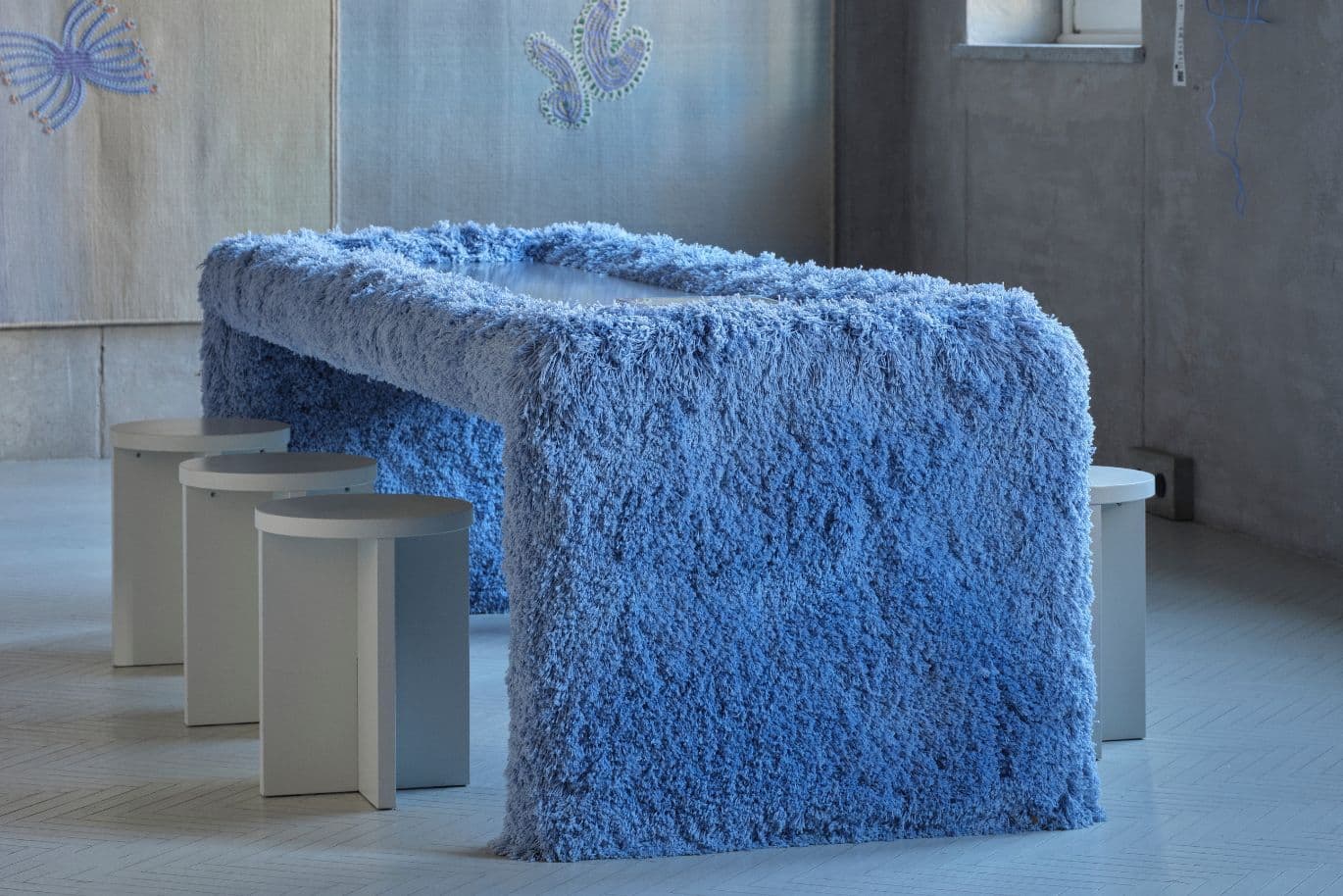 Ein moderner rechteckiger Tisch, der mit blauem, zotteligem Stoff bezogen ist, umgeben von minimalistischen weißen Hockern in einem Betonraum.