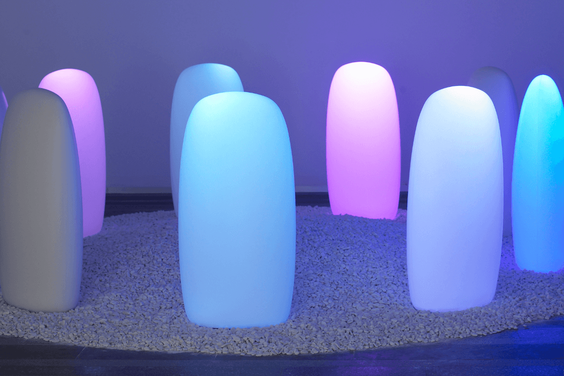 Bunte, säulenförmige Lichter, die in Blau- und Rosatönen auf einem Bett aus weißen Kieselsteinen in einem dunklen Raum leuchten.