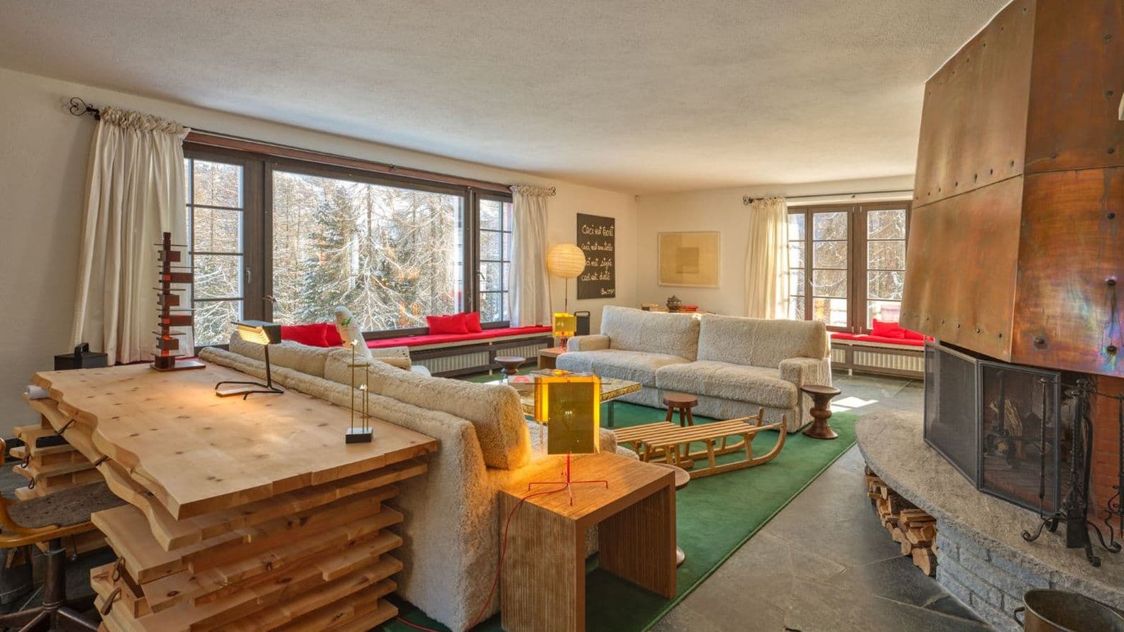 Sonnenbeschienener Wohnraum mit weichen cremefarbenen Sofas, grünem Teppich, gestapeltem Holztisch, Schlitten, großen Fenstern mit Blick auf verschneite Bäume und einem Kupferkamin.