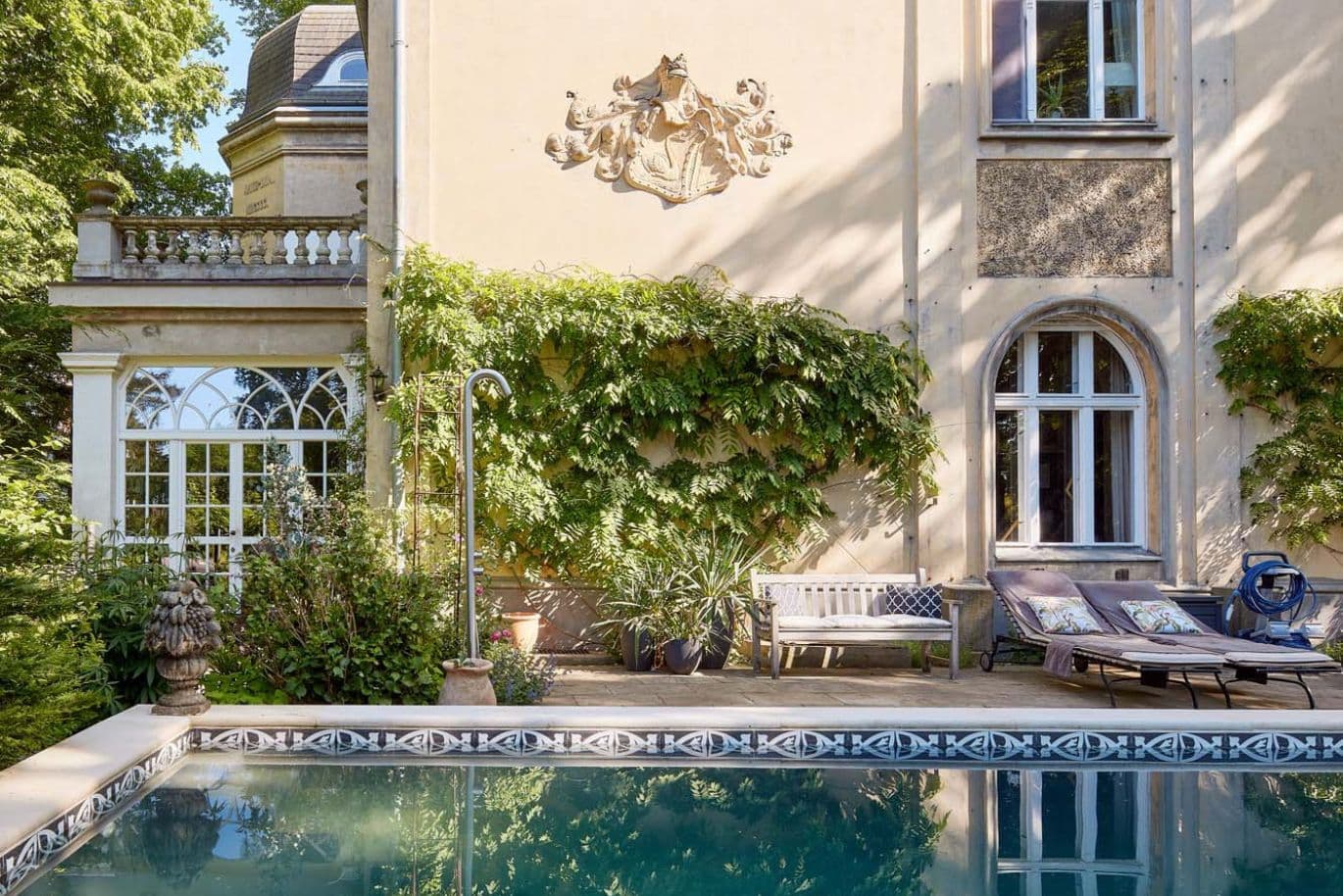 Elegant villa with an ornate façade, lush greenery, and a tranquil pool reflecting the building. Sunlit trees frame the scene beneath a clear blue sky.