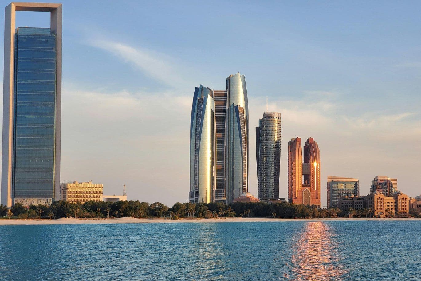A skyline of modern skyscrapers, including reflective towers, stands along the waterfront under a clear blue sky.