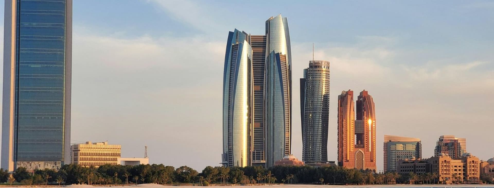 A skyline of modern skyscrapers, including reflective towers, stands along the waterfront under a clear blue sky.