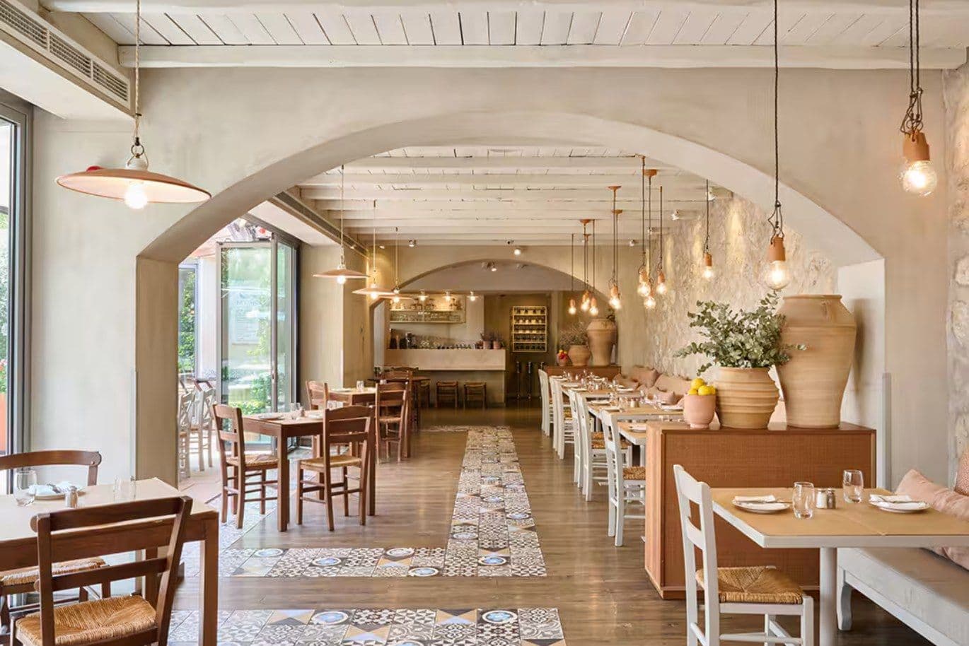 Cozy restaurant interior with wooden tables and chairs, decorative tiles, hanging lights, large vases, and a view of the outdoor area through glass doors.