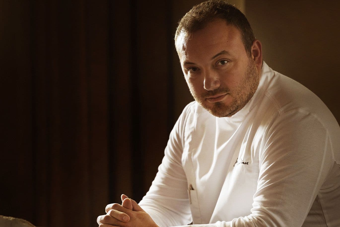 A chef seated in a warmly lit room, wearing a white jacket and resting his hands on the table, looking thoughtfully at the camera.