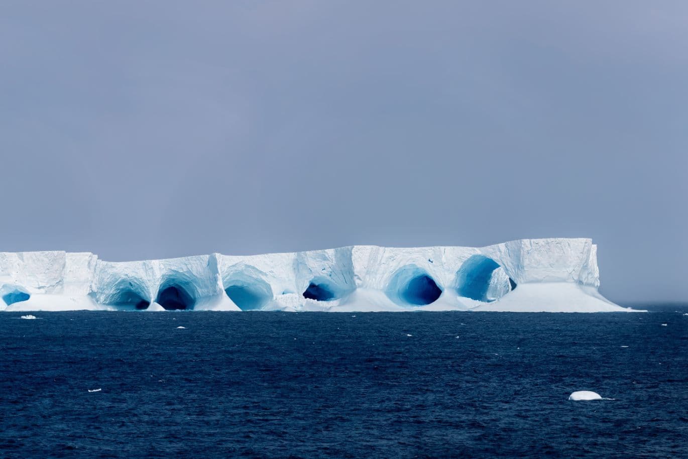 Ein Eisberg mit glatten Bögen und Tunneln, der unter einem bewölkten Himmel in einem ruhigen blauen Ozean schwimmt.