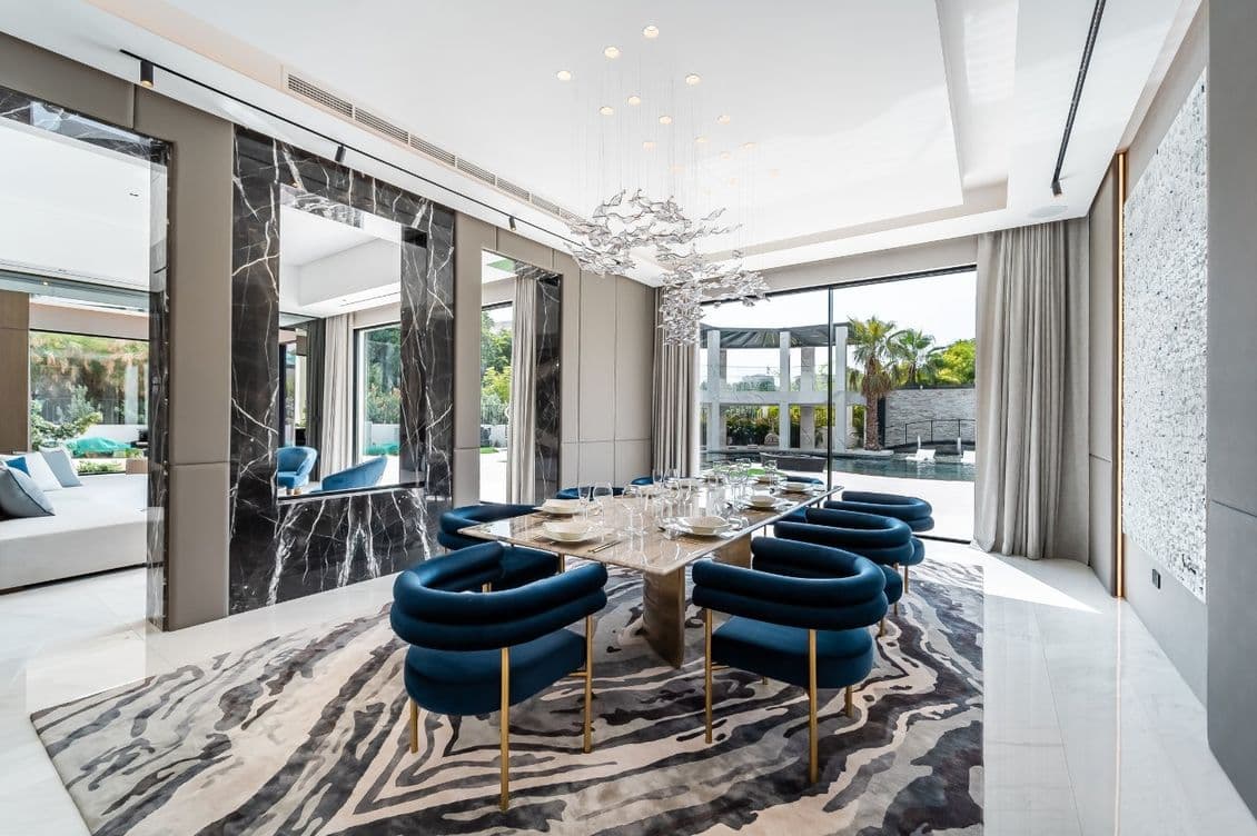 Modern dining room with a large glass table, blue chairs, abstract chandelier, marble accents, and a view of a patio through large windows.