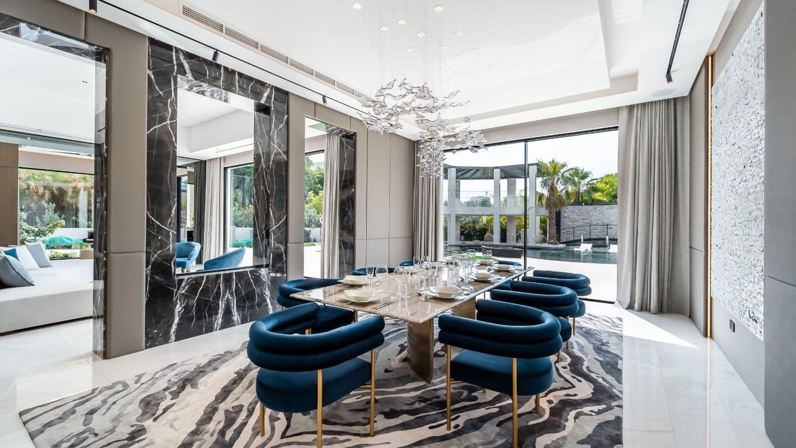 Modern dining room with a large glass table, blue chairs, abstract chandelier, marble accents, and a view of a patio through large windows.