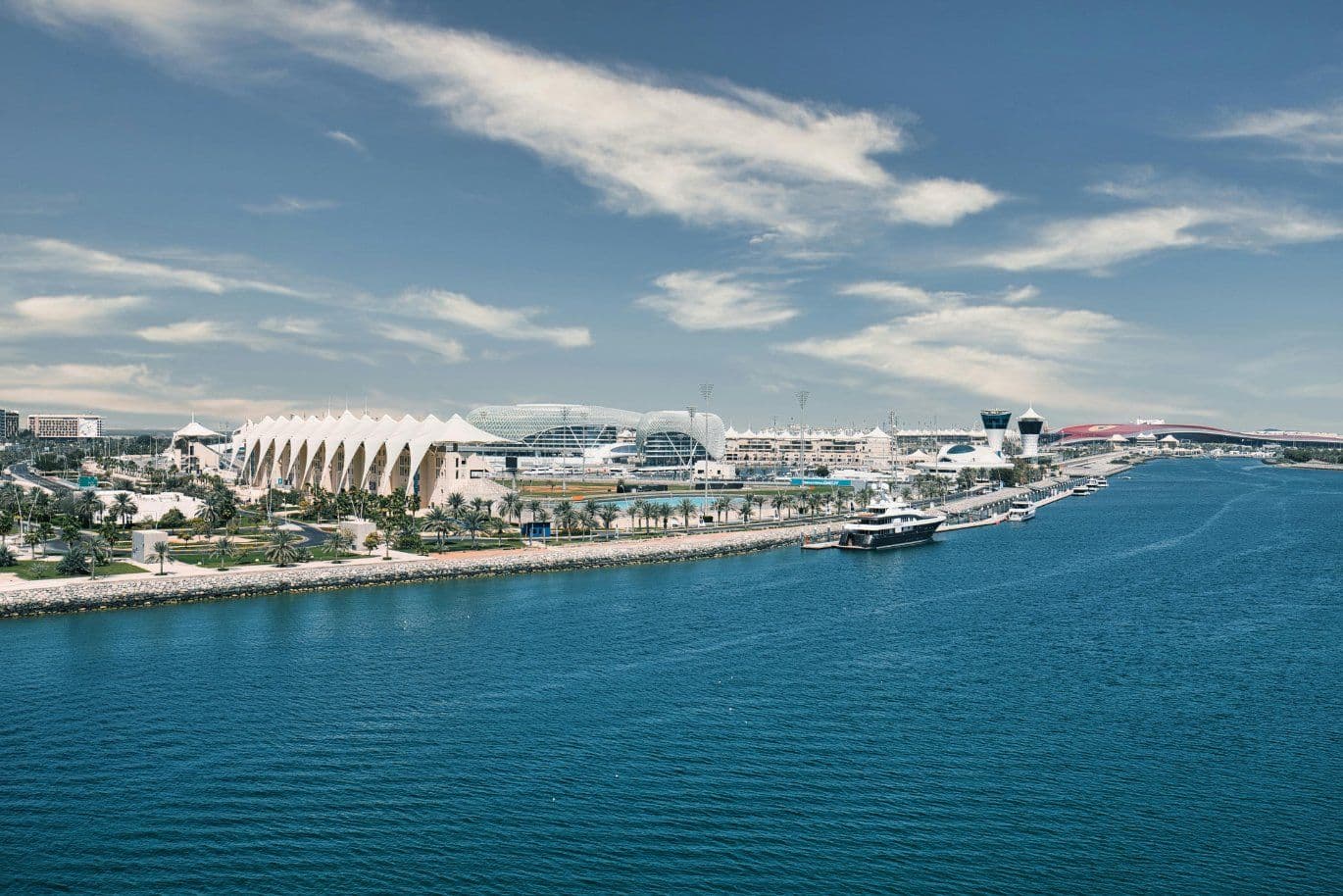 Aerial view of a waterfront with modern architecture and a marina, featuring a unique building and several yachts under a partly cloudy sky.