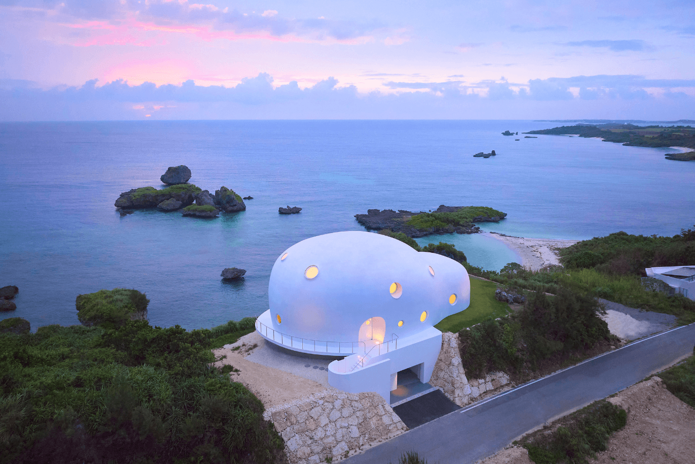 Ein futuristisches weißes Kuppelhaus mit runden Fenstern an einer felsigen Küste, vor dem Hintergrund eines farbenprächtigen Sonnenuntergangs über dem Meer.
