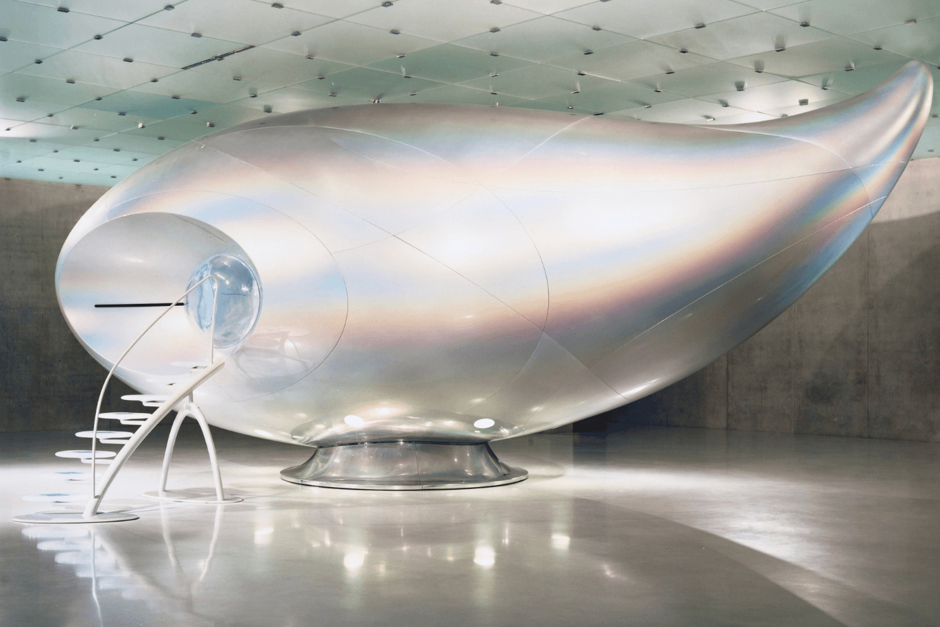 Eine futuristische, kapselartige Struktur mit schillernder Oberfläche und einer kleinen weißen Treppe, die zum Eingang führt, eingebettet in einen modernen, minimalistischen Raum.