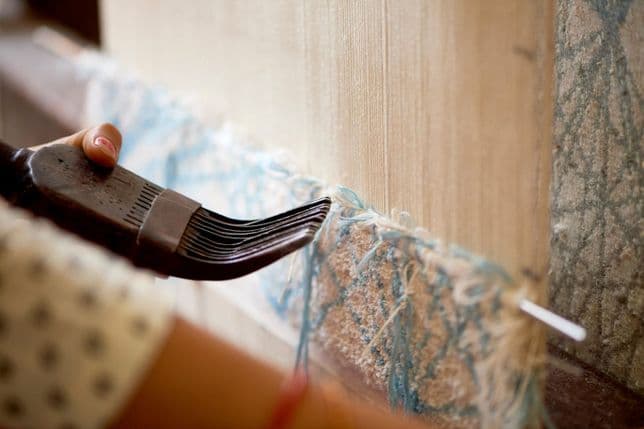 Nahaufnahme einer Hand, die mit einem traditionellen Kammwerkzeug ein teilweise gewebtes blau-weißes Textil kämmt.