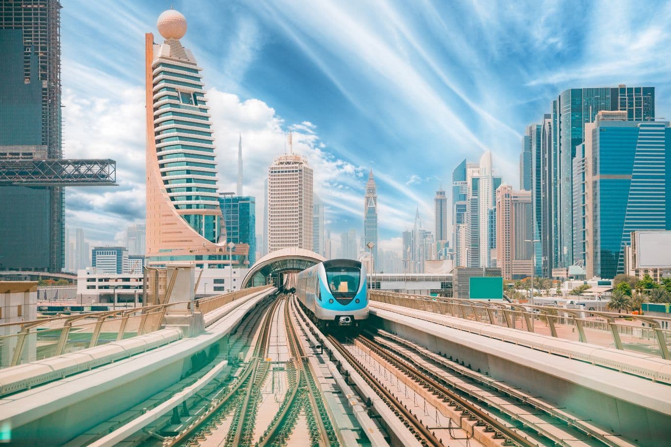 A modern train travels on elevated tracks through Dubai with futuristic skyscrapers and a bright, cloudy sky.