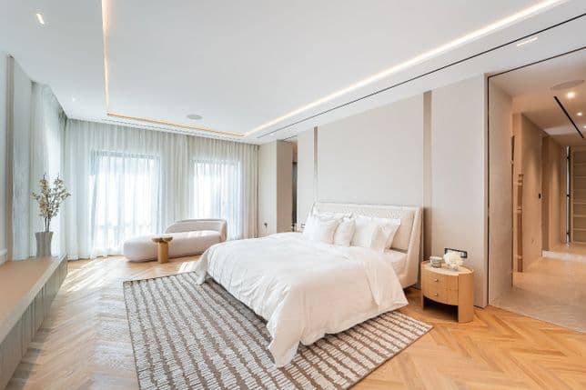 Modern bedroom with a large bed, white bedding, patterned rug, cushioned chair, and floor-to-ceiling windows with sheer curtains.