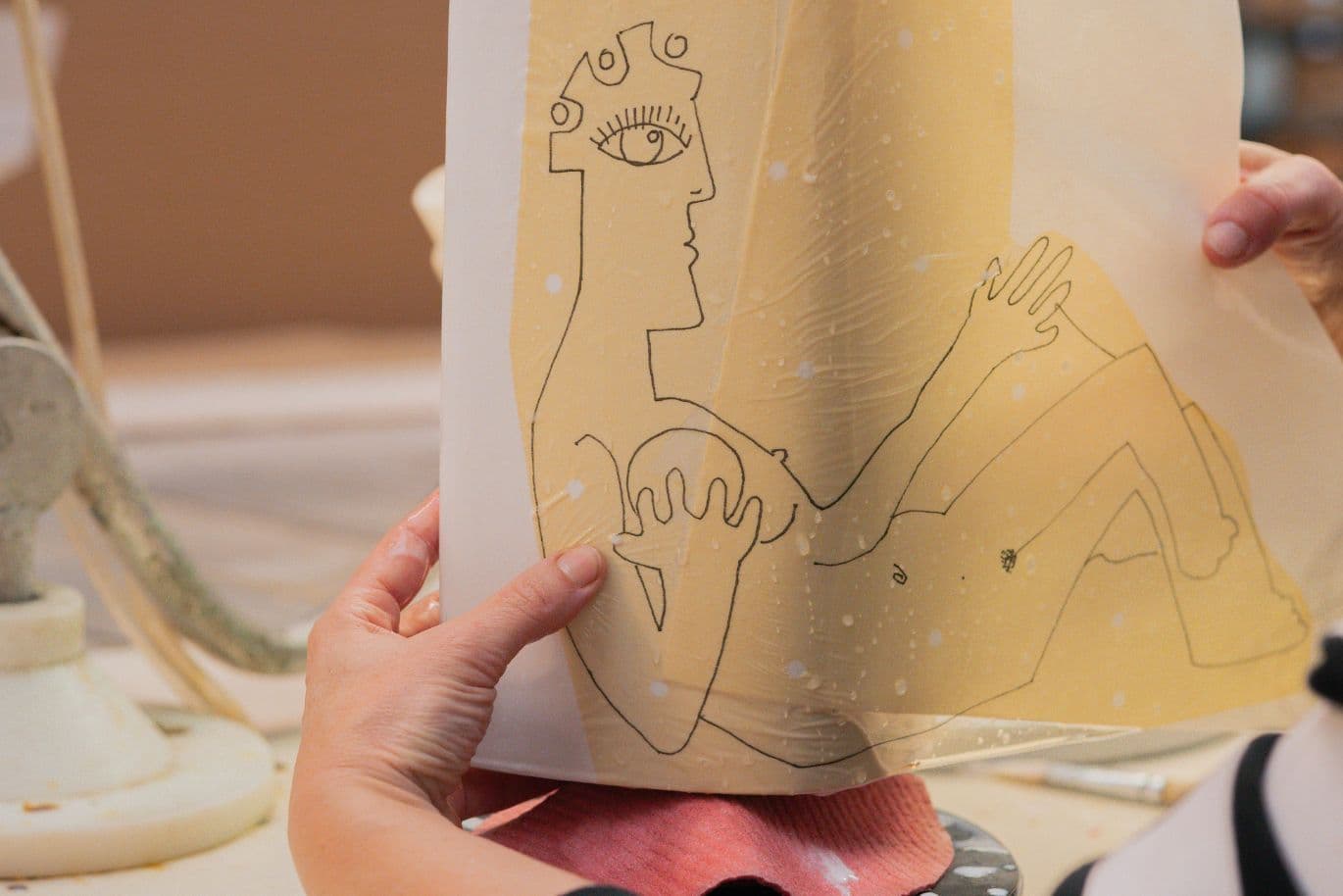 Hände, die ein durchscheinendes Blatt mit einer minimalistischen Zeichnung einer liegenden Figur halten, umgeben von Kunstmaterialien.