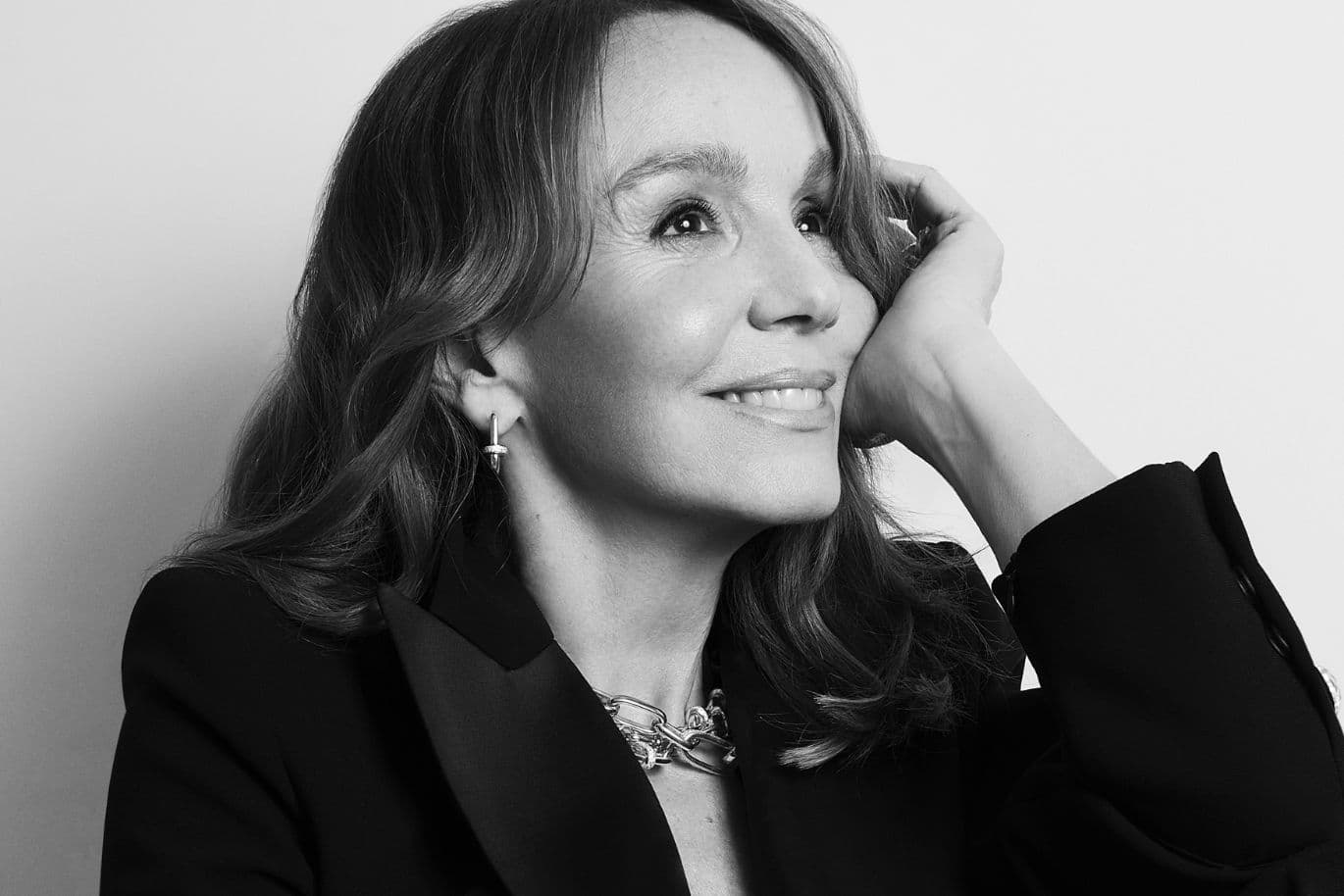 Black-and-white portrait of a smiling woman with shoulder-length hair, wearing a blazer and necklace, resting her head on her hand.