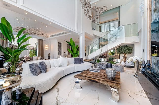 Luxurious, spacious living room with modern decor, white marble floors, a curved sofa, large plants, a glass staircase, and high ceilings.