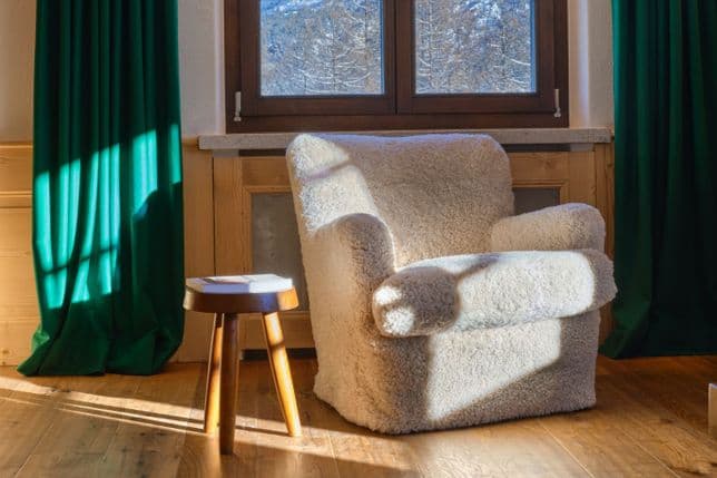 Plüschiger, cremefarbener Schaffell-Sessel, der vom Sonnenlicht durchflutet wird, neben einem Fenster mit dunkelgrünen Vorhängen, daneben ein kleiner Holzhocker mit einem Buch.