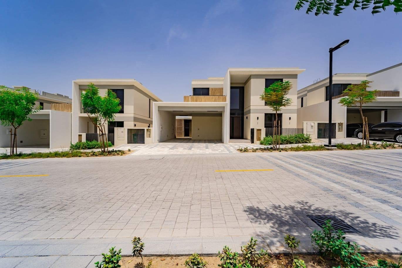 Modern suburban house with a flat roof, large windows, and a driveway, surrounded by small trees and landscaped greenery.