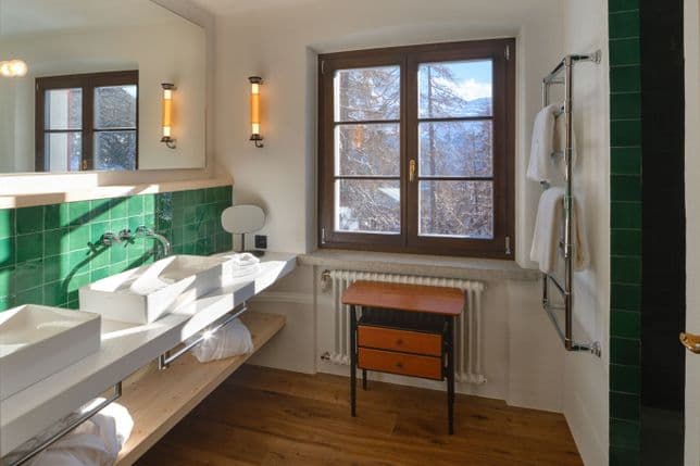 Helles Badezimmer mit grüner Fliesenrückwand, zwei weißen Waschbecken, Holzfensterrahmen mit Blick auf verschneite Bäume, kleinem Holztisch und verchromtem Handtuchhalter.