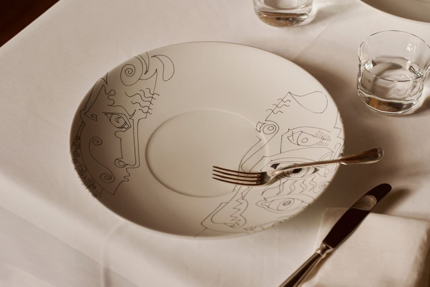 Weißer Teller mit abstrakten Gesichtsdesigns, auf einem Tisch mit Gabel, Messer und Gläsern platziert. Schlichte und elegante Tischdekoration.